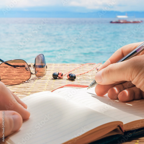 caucasian man is writing some idea, message or letter to his notepad by pen while he sitting on the beach of tropical sea with boat