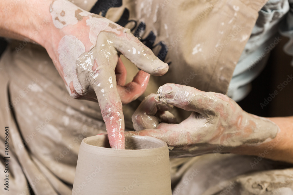 pottery, workshop, ceramics art concept - top view on man's hands work with potter wheel and raw clay, male fingers with water sculpt some new jug, close-up