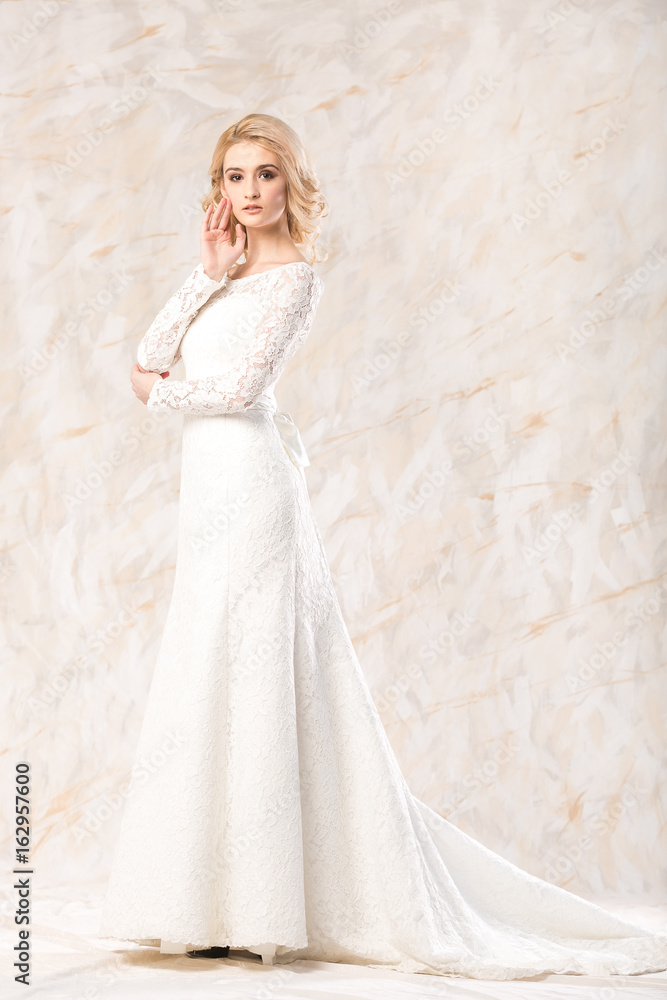 fashionable white gown, beautiful blonde model, bride hairstyle and makeup concept - young charming lady in wedding festive dress with train standing indoors on light background, pretty woman posing