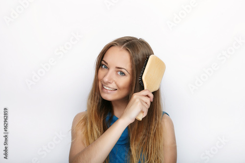 Headshot of young adorable blonde woman with cute smile with comb in hand brush her hair on white background