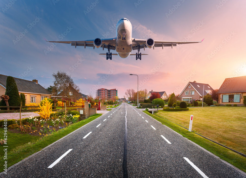 Fototapeta premium Beautiful cityscape with passenger airplane is flying in the sunset sky above the asphalt road through the town with houses and courtyards at sunset in Netherlands. Landing airplane. White aircraft