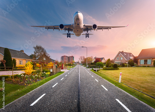 Beautiful cityscape with passenger airplane is flying in the sunset sky above the asphalt road through the town with houses and courtyards at sunset in Netherlands. Landing airplane. White aircraft