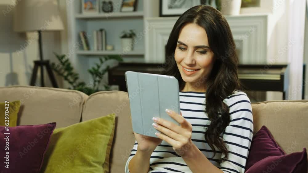Happy beautiful woman using tablet device, reading watching video and smiling. Sitting on the couch in the living room. Close up Happy beautiful woman using tablet device, reading watching video and smiling. Sitting on the couch in the living room. Close up