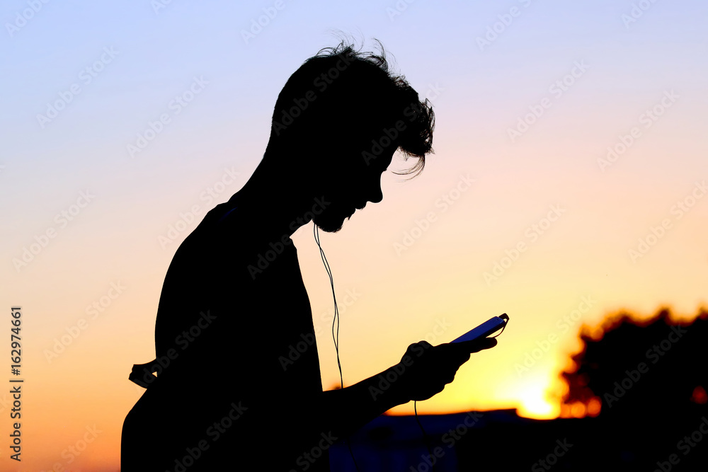 silhouette of a man with a mobile phone and headphones at sunset.