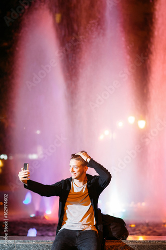 Young man take selfie on smartphone while walking in night city streets. Smiling guy on blurred lights background