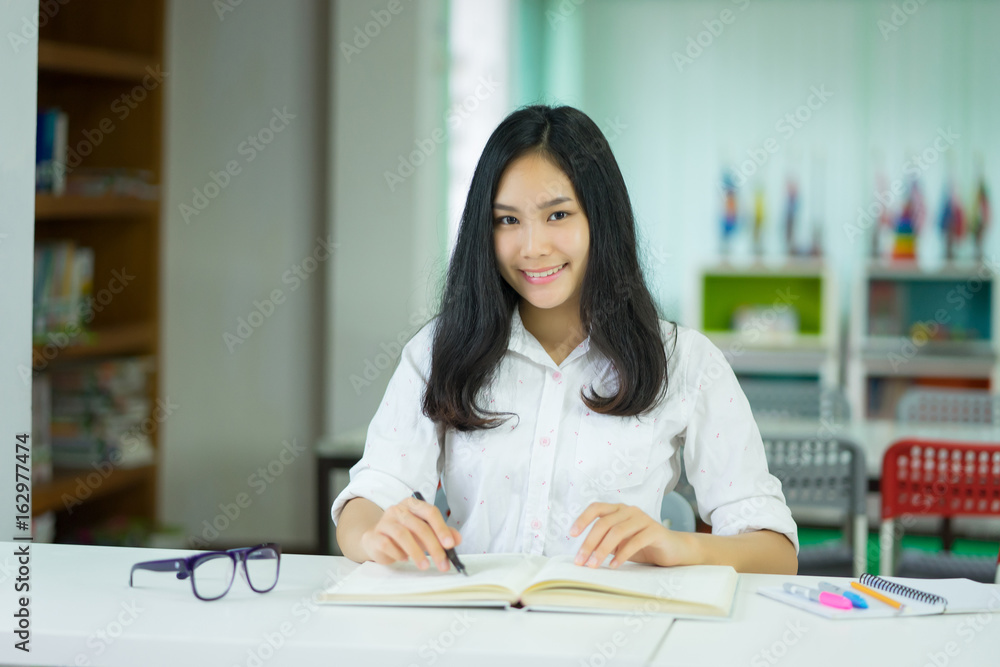 Female student taking notes preparing exam and learning lessons from a book at library. Young asian woman sitting at table doing assignments making research look at camera ,smiling in college librarys Female student taking notes preparing exam and learning lessons from a book at library. Young asian woman sitting at table doing assignments making research look at camera ,smiling in college librarys