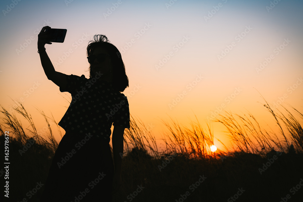 Silhouette of woman taking selfie during sunset with flowering grass