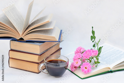 Beautiful pink flowers are in the open book near stack of other books and coffee mugs.
