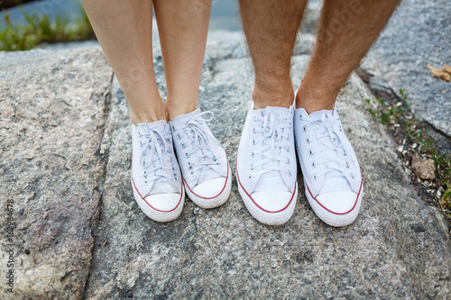 Love story told by boots. Human feet close up. Man and woman in sneakers. Girl in white shoes. Guy in black sneakers and denim. Hipster couple in summer. Legs close up.