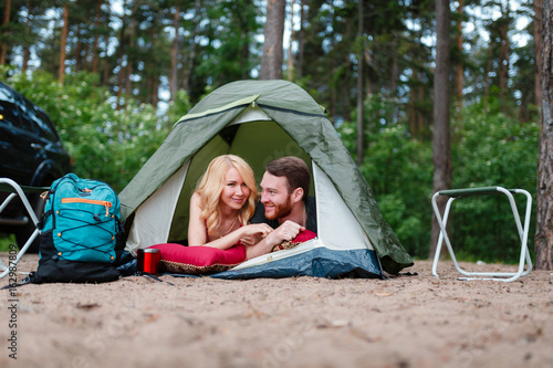 Beautiful couple lying in a tent, camping on the background of fire. Next to folding chairs, camping a mug of warm soup and a Hiking backpack. Beside their off-road machine.