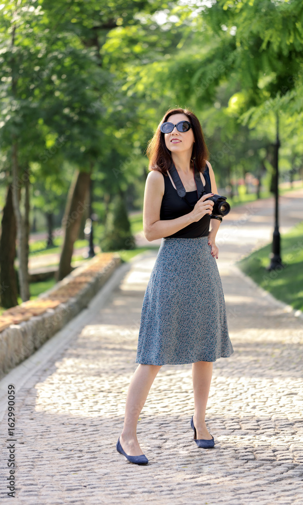 Young woman (photographer, tourist) in a blue skirt and black top holds a camera in her hand. Young woman (photographer, tourist) in a blue skirt and black top holds a camera in her hand.