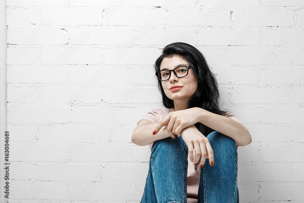 Portrait of smart beautiful brunette girl in eyeglasses with natural make-up sitting near white brick wall. Portrait of smart beautiful brunette girl in eyeglasses with natural make-up sitting near white brick wall.