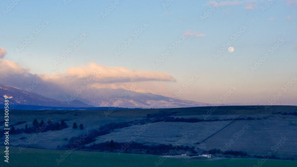 Spring foothills with snowy and cloudy high mountains at twilight.