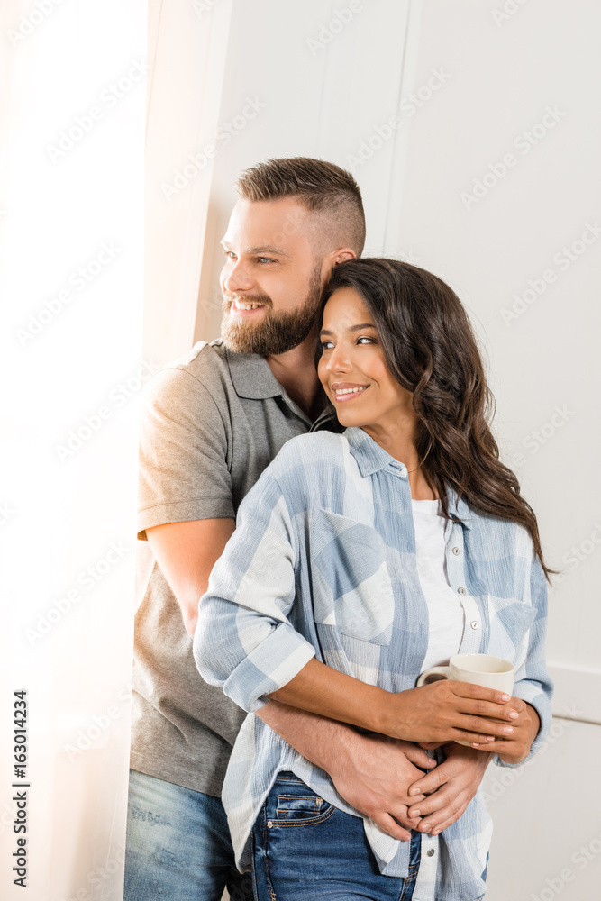 Happy young couple standing embracing and looking at window at home Happy young couple standing embracing and looking at window at home