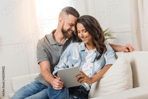 tender smiling couple with digital tablet sitting on sofa at home