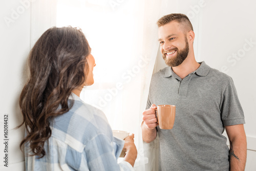 Happy young couple holding tea cups and smiling each other at home