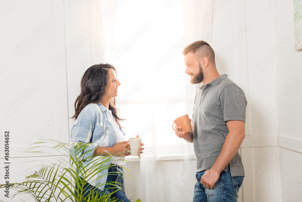 Side view of cheerful young couple holding tea cups and smiling each other at home Side view of cheerful young couple holding tea cups and smiling each other at home