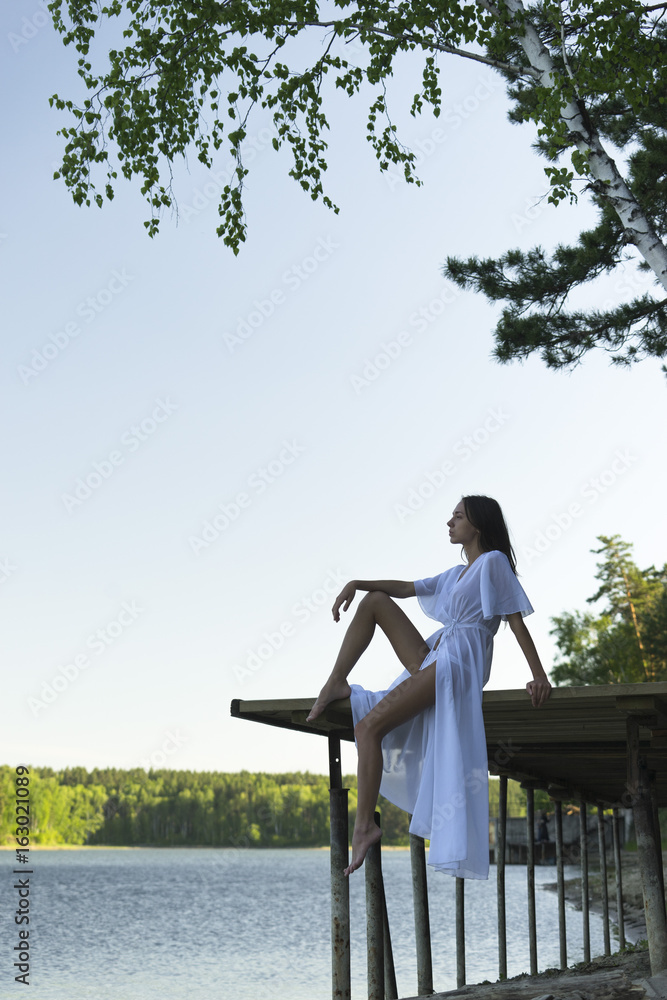 Young beautiful brunette woman in white dress sits on the wooden bridge near forest river Young beautiful brunette woman in white dress sits on the wooden bridge near forest river