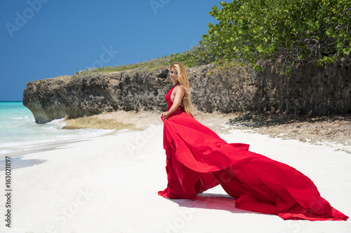 Beautiful young woman with blond long hair in red fluttering dress stands aside on the coastline of azure caribbean sea