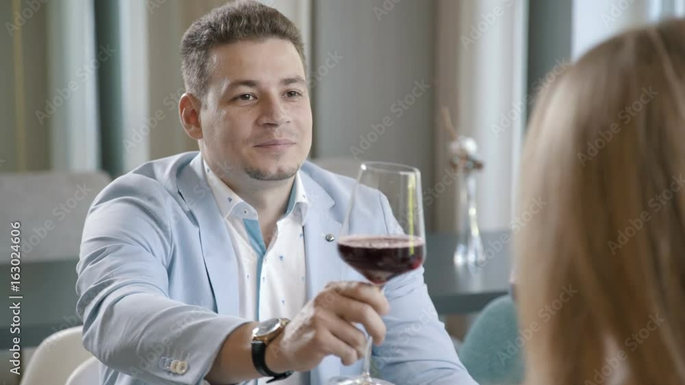 Young man in a stylish light business sits on a light background with a glass of red wine and speaks a toast. A stylish businessman in a restaurant with his wife: a romantic dinner. Young man in a stylish light business sits on a light background with a glass of red wine and speaks a toast. A stylish businessman in a restaurant with his wife: a romantic dinner.