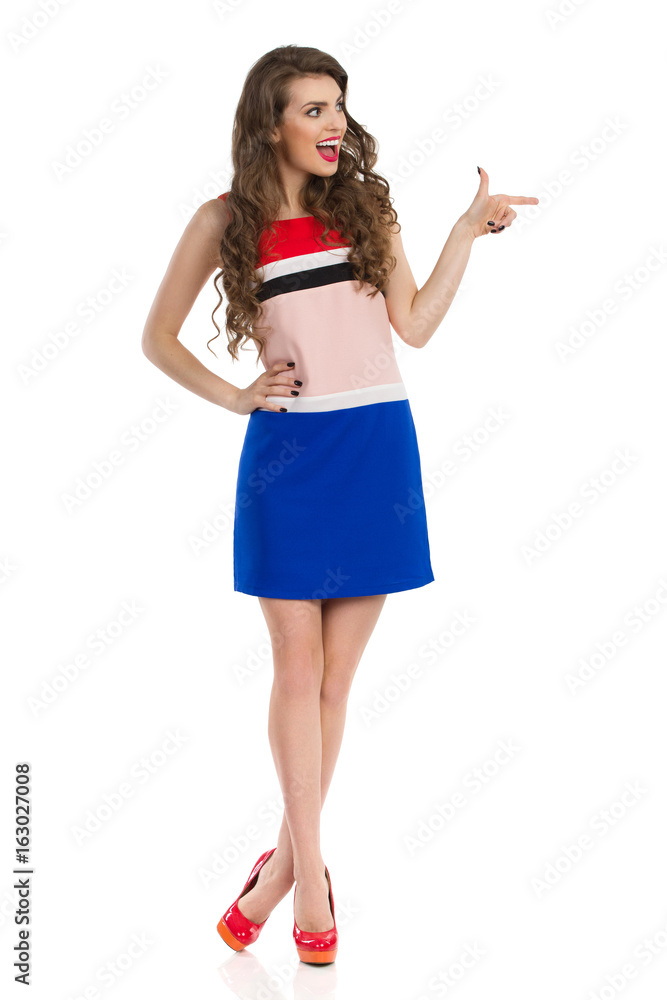 Fototapeta premium Excited Woman In Colorful Mini Dress And High Heels Is Looking Away And Pointing