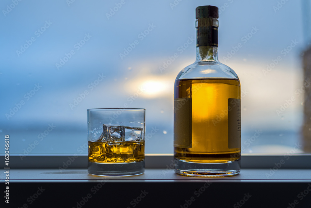 Amber colored alcohol drink with bottle on rainy window sill Amber colored alcohol drink with bottle on rainy window sill