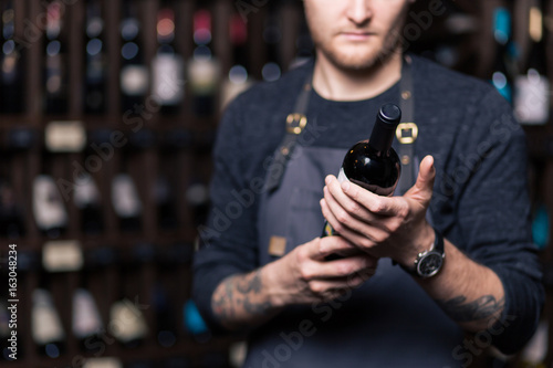Focused young male sommelier in suite looking at red wine in bottle photo