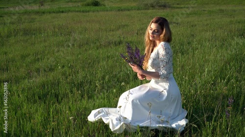Beautiful young woman sitting on the meadow with flowers on the sunset