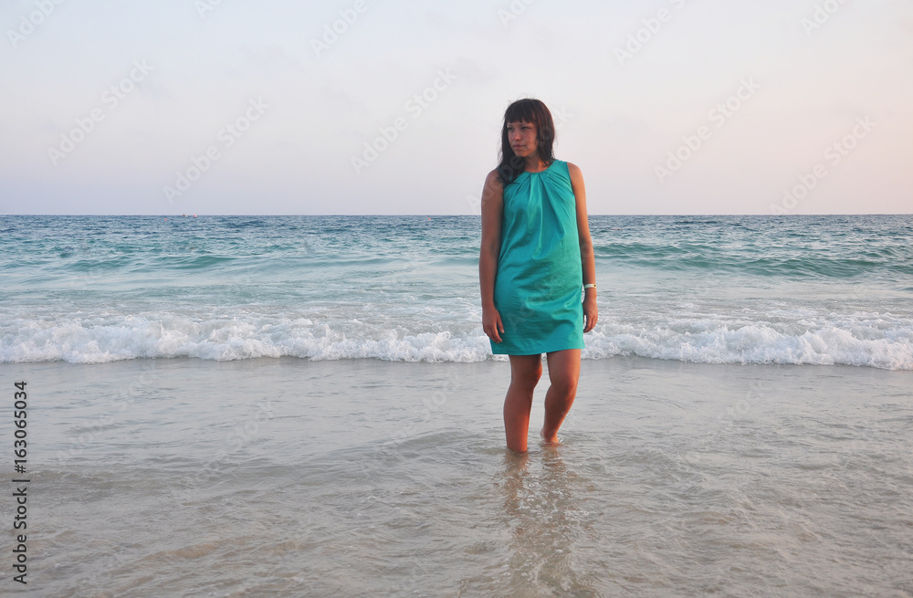 beautiful brunette girl in a short green sundress with a smooth southern sun on the sea background. legs washed by the surf. evening on the beach