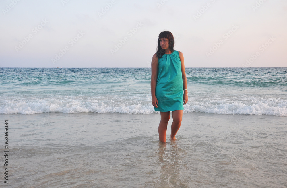 beautiful brunette girl in a short green sundress with a smooth southern sun on the sea background. legs washed by the surf. evening on the beach beautiful brunette girl in a short green sundress with a smooth southern sun on the sea background. legs washed by the surf. evening on the beach