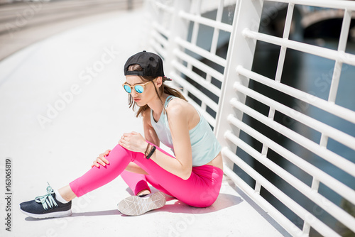 Young woman having an injury and pain in her knee sitting outdoors on the white bridge