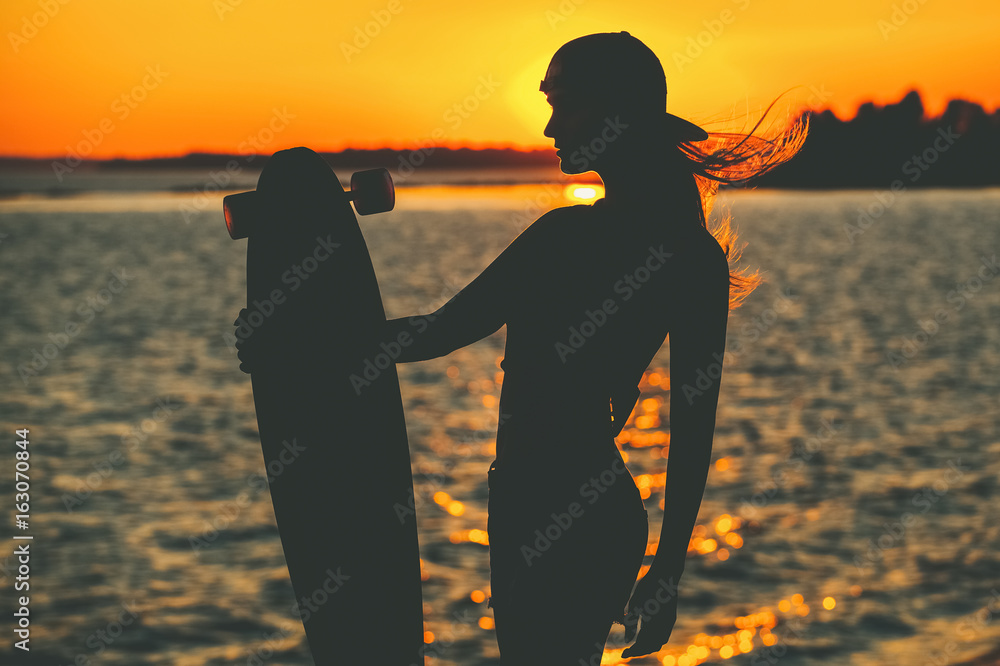 Photo & Art Print Silhouette of a beautiful, sexy hipster girl in tattoos  standing with a longboard against the sunset at the sea