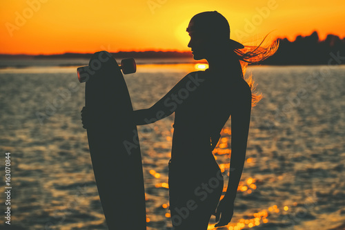 Canvas Print Silhouette of a beautiful, sexy hipster girl in tattoos standing with a longboard against the sunset at the sea
