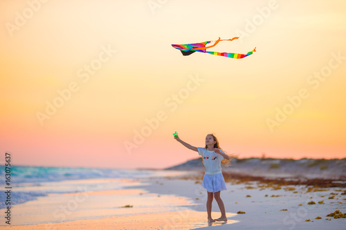 Little girl with flying kite on tropical beach at sunset. Kid play on ocean shore. Child with beach toys.