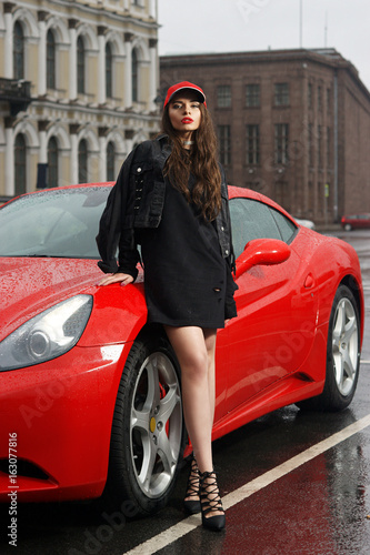 Glamorous sexy fashionable woman wearing sweetshot, jacket and red cap with long brown hair standing against red sport car at city street on overcast day © Dmitry Tsvetkov