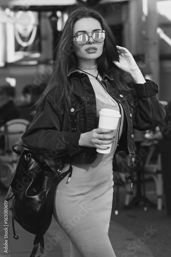 Young beautiful stylish woman wearing green dress, black jeans jacket and sunglasses walking at the street in city. Pretty brunette girl with long hair holding backpack and coffee