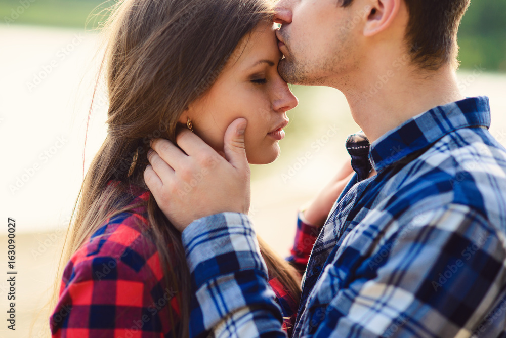 Stylish young couple in shirts and jeans while walking along the lake shore. A handsome boyfriend kisses his beautiful charming girlfriend on the forehead. Close up. Stylish young couple in shirts and jeans while walking along the lake shore. A handsome boyfriend kisses his beautiful charming girlfriend on the forehead. Close up.