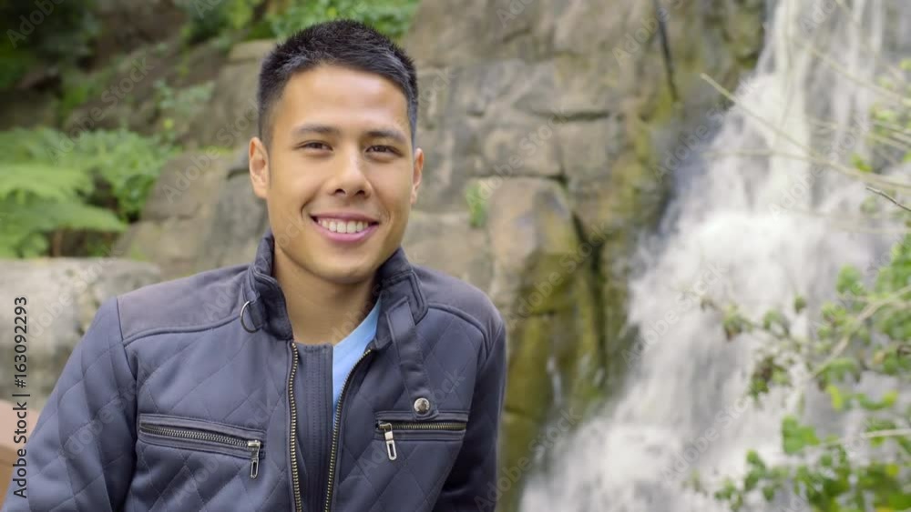 Man Stands In Front Of Waterfall, Looks Around At Scenery, Smiles At Camera Man Stands In Front Of Waterfall, Looks Around At Scenery, Smiles At Camera