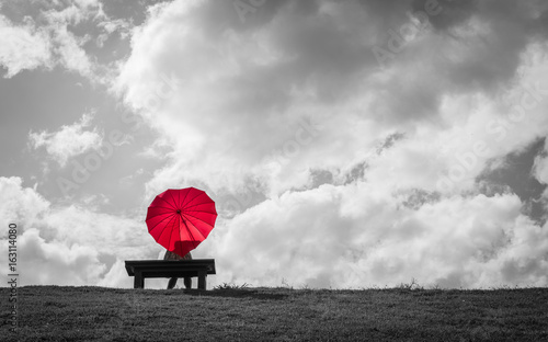 a women sitting alone on a bench with a red heart shaped umbrella waiting for love
