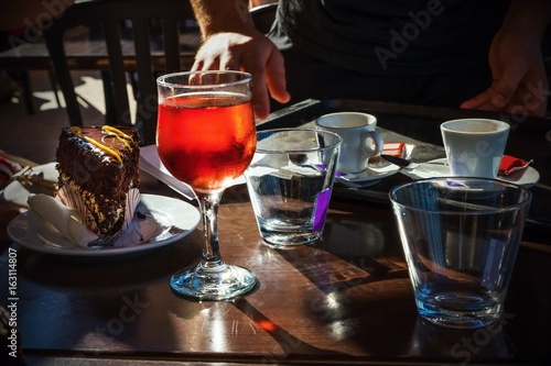 Table, light glass of red beverage, coffe, cake, water, glass, hand. Dark