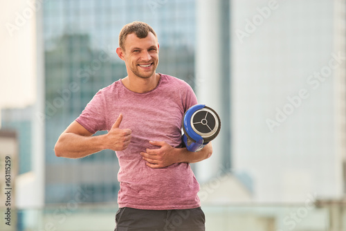 Man holding hoverboard and smiling. Guy showing thumb up. Make your trips easy.