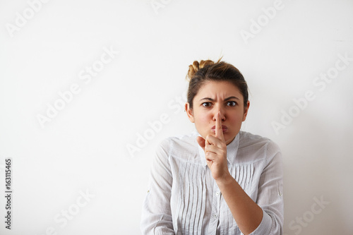Emotional funny student girl or employee keeping index finger at her lips, asking to keep confidential information a secret, standing at blank wall with copy space for your promotional content