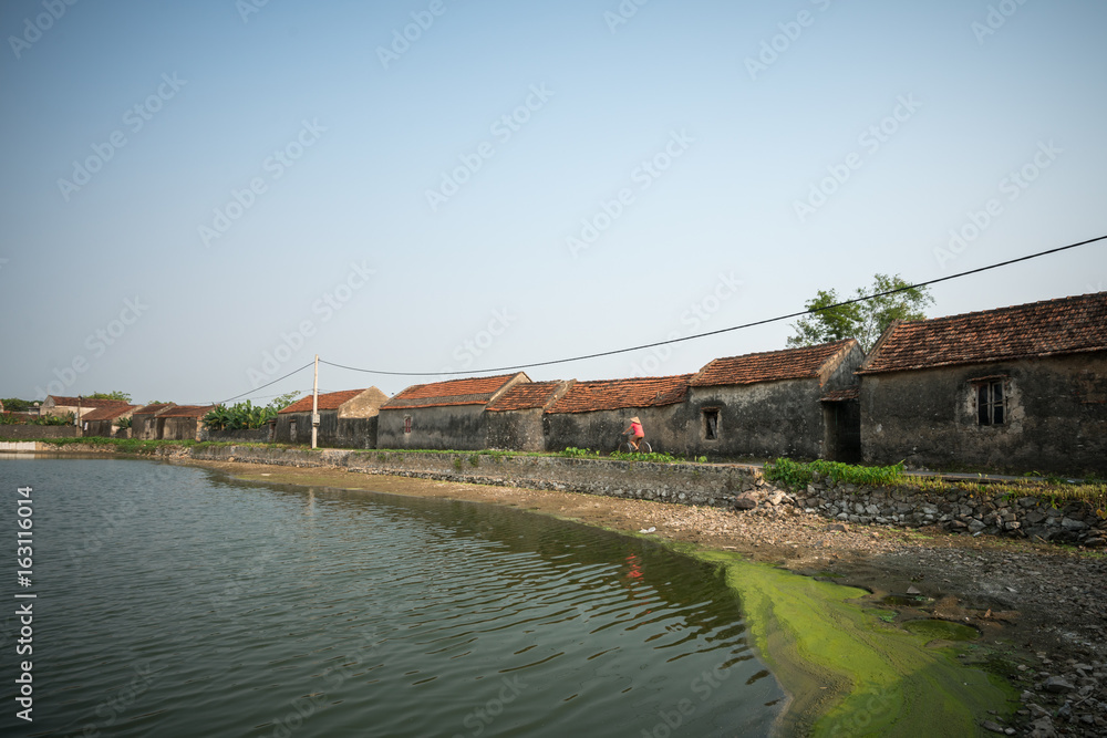 Vietnam landscape with pond and old aged houses and woman cycling on road in village