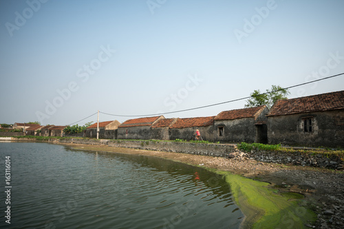Vietnam landscape with pond and old aged houses and woman cycling on road in village
