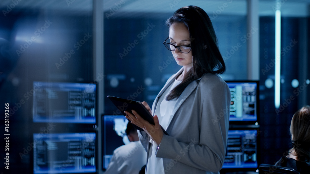 Young Female Engineer Uses Tablet in System Control Center. In the Background Her Coworkers are at Their Workspaces with many Displays Showing Valuable Data. Young Female Engineer Uses Tablet in System Control Center. In the Background Her Coworkers are at Their Workspaces with many Displays Showing Valuable Data.