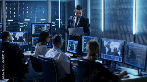Professional IT Engineers Working in System Control Center Full of Monitors and Servers. Supervisor Holds Laptop and Holds a Briefing. Possibly Government Agency Conducts Investigation. photo