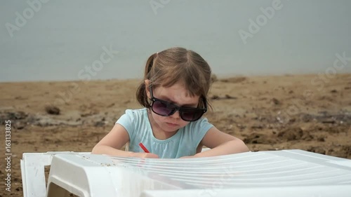 A frustrated girl in big sunglasses. She sits on the seashore, draws. photo