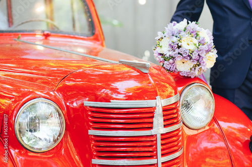 Fotografie The groom is holding a wedding bouquet on the hood of a red retro car