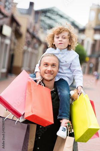 portrait of smiling father piggybacking his little son with shopping bags at street