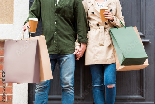 cropped shot of casual couple standing with shopping bags and drinking coffee on street